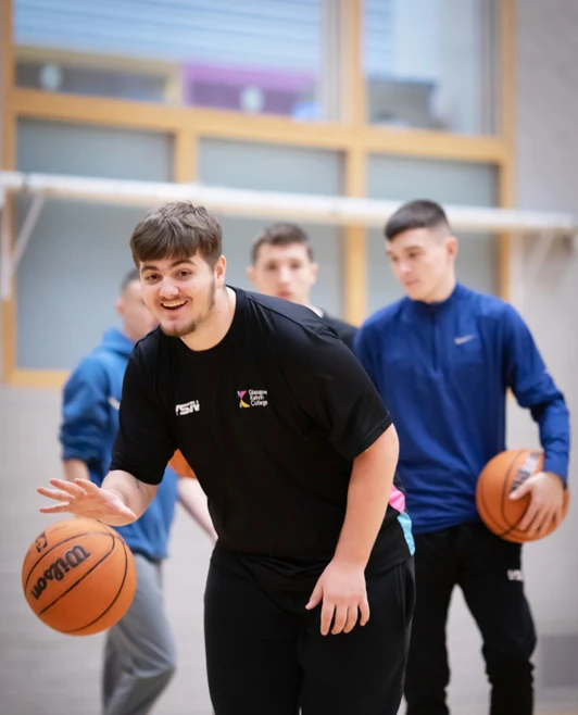 A student dribbling a basketball indoors, smiling, with other students and basketballs in the background. A student dribbling a basketball indoors, smiling, with other students and basketballs in the background.
